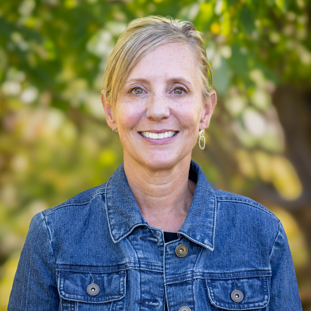 A woman with blond hair is wearing a jean jacket and smiling at the camera