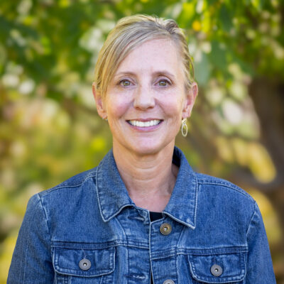 A woman with blond hair is wearing a jean jacket and smiling at the camera