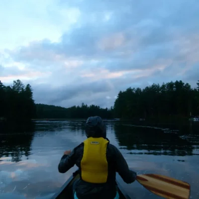 Student wearing yellow personal floatation device paddles a canoe.