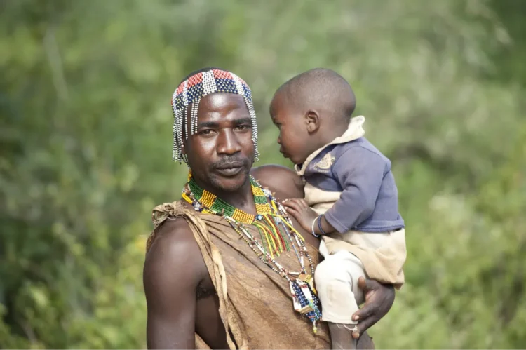 A Tanzanian father wearing traditional ornaments carries his son.