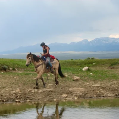 Horse packing student rides along the water's edge with mountains in the background.