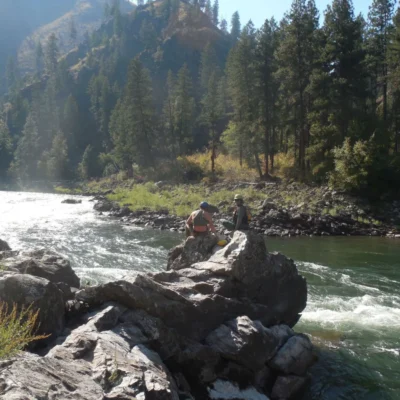Two students take a break on a rocky riverbank, with white water rushing past them and tall pine trees covering the forested hillside beyond.
