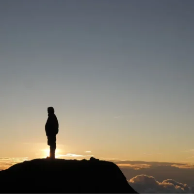 Silhouetted figure stands at the top of a peak in Tanzania with sun illuminating the clouds that hug the mountains.