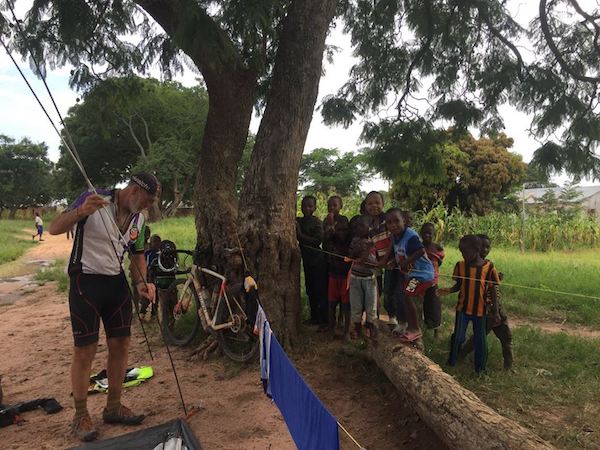 TDA cyclist on a bike tour pitches a tent near clothesline while group of children watches
