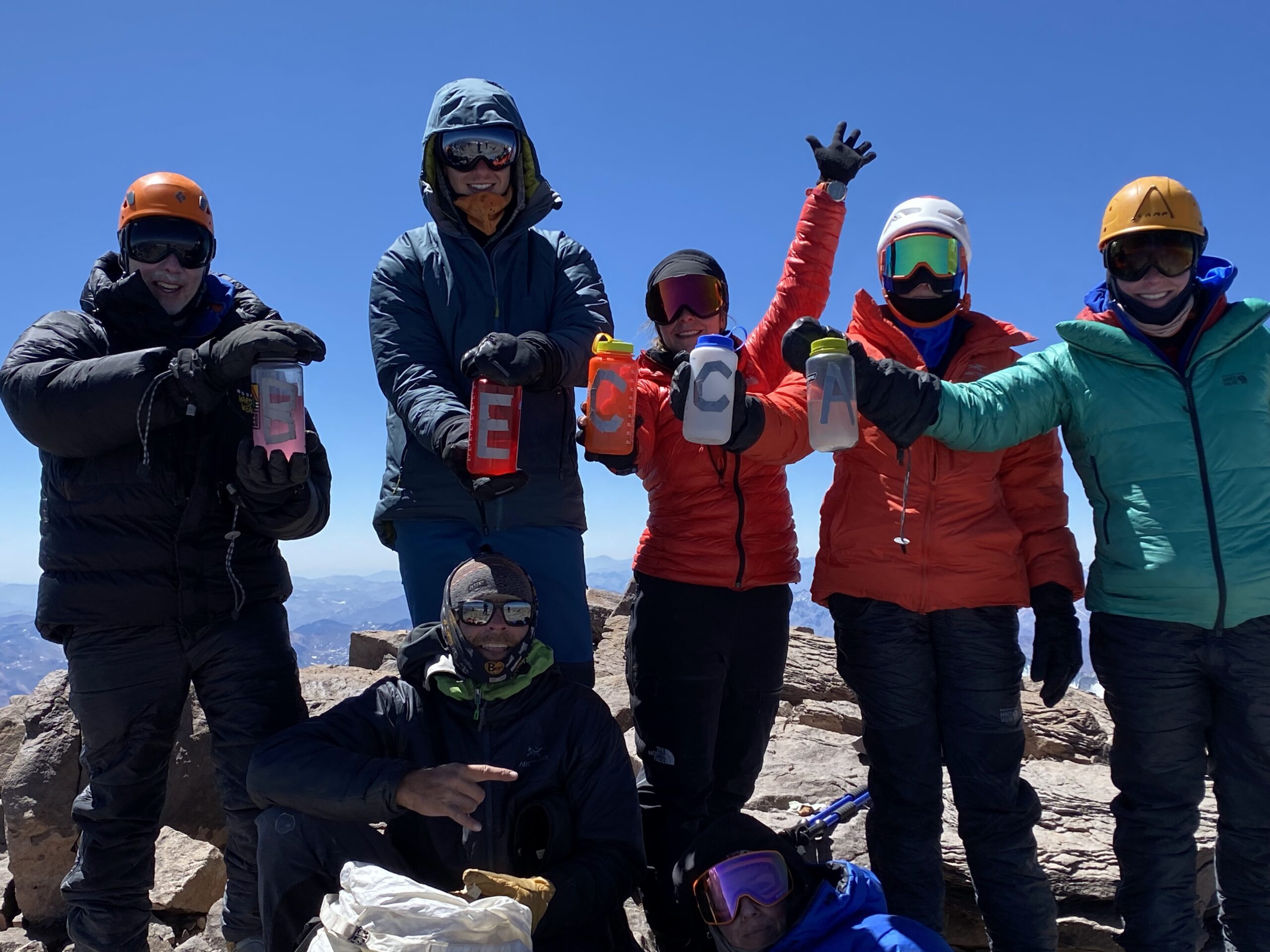 Six people standing at the summit of Aconcagua holding up water bottles with letters to spell: BECCA