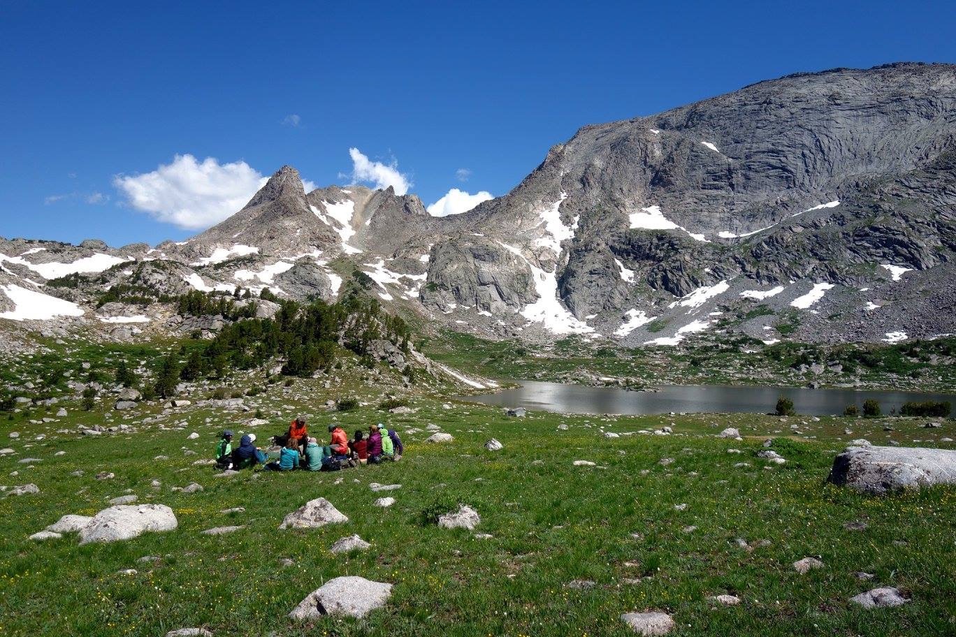 Group of NOLS students sits in a circle beside an alpine lake