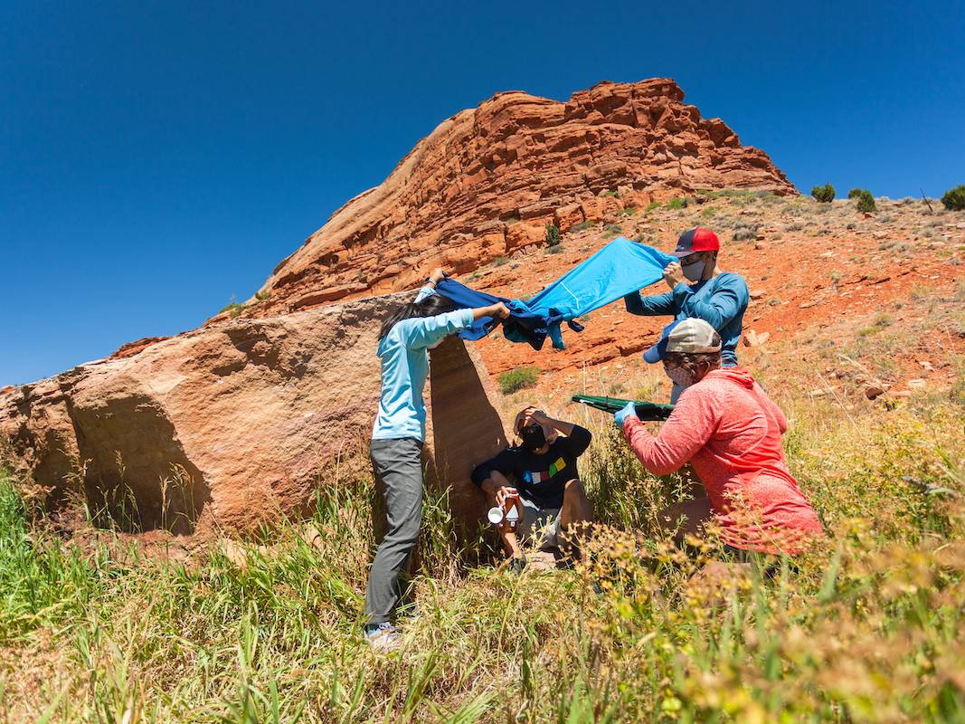 Three people shade and fan an individual sitting on the ground in a hot, sunny field