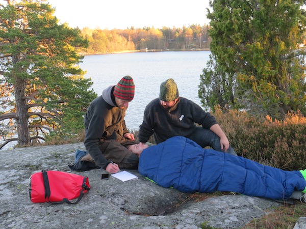 two NOLS Wilderness Medicine students practice caring for a patient lying in sleeping bag with lake and trees behind