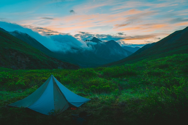NOLS tent pitched in lush green valley in Alaska with sunset over mountains draped in clouds
