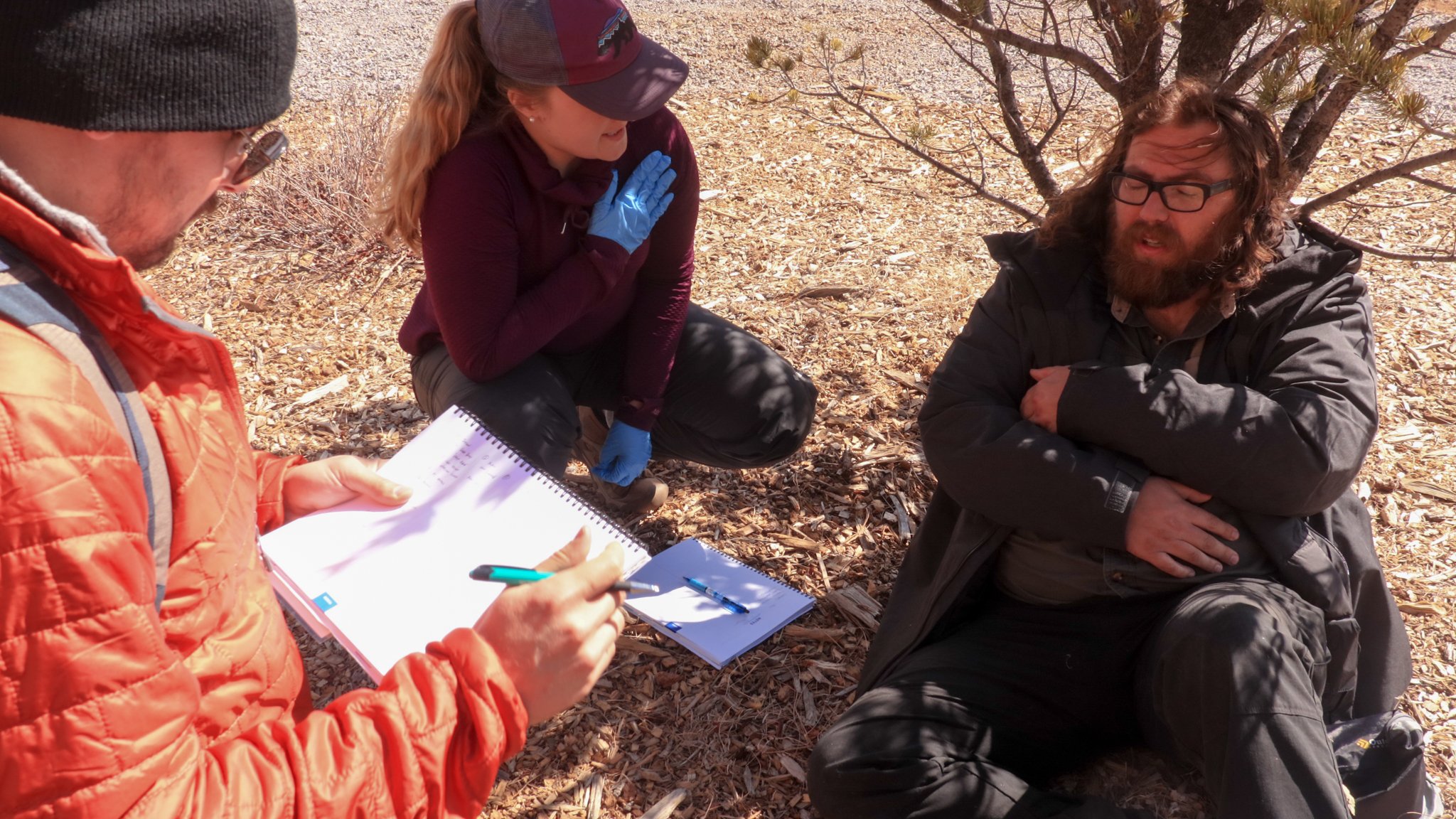 Two rescuers, one male, one female, approach a male patient, who clutches his chest and sits under a tree. A male rescuer takes notes. 