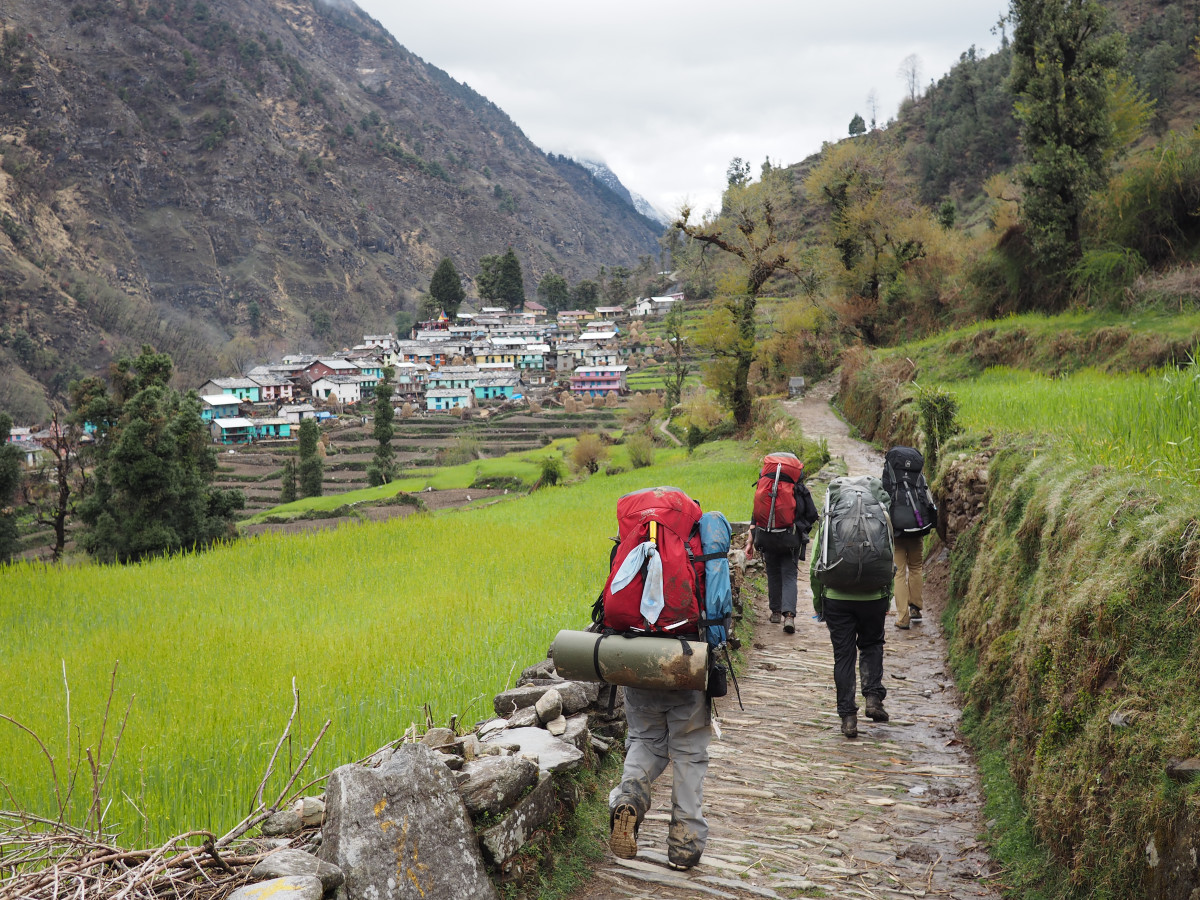 NOLS students hike wearing backpacks on a path in India with colorful village buildings in the background