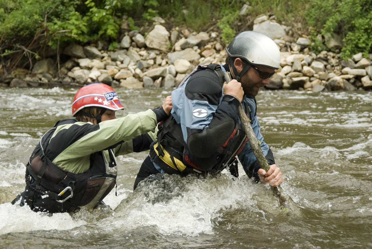 Two people cross a river using a large stick for assistance