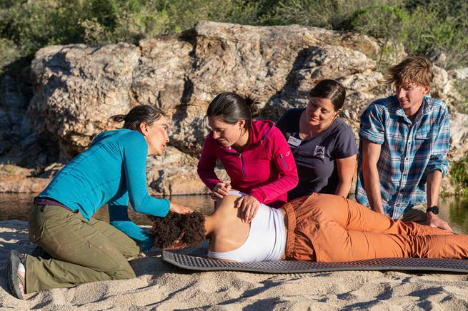 A NOLS instructor instructing 3 people on how to properly care for an injured person in the wilderness, with a 4th person laying and pretending to be injured