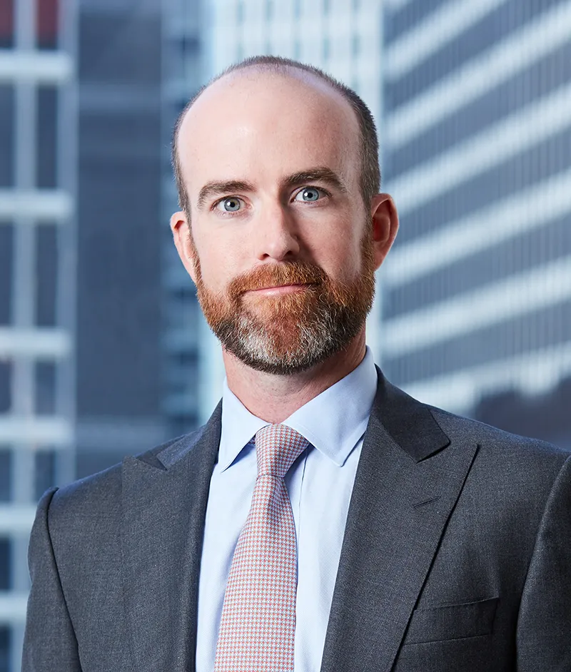 A man with a suit jacket and tie stands against a skyscraper background. He has a red mustache and beard.