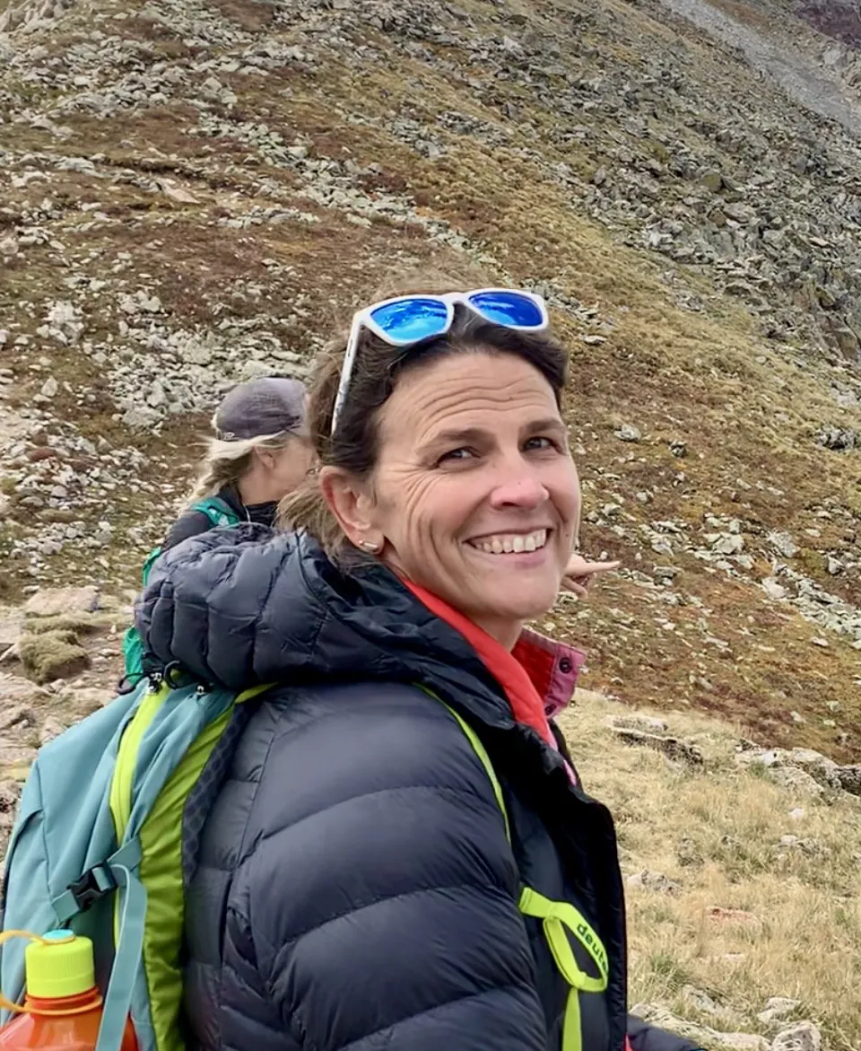 A woman with brown hair and blue sunglasses stands against a grassy mountainside. She is wearing a blue puffy jacket and is smiling at the camera.