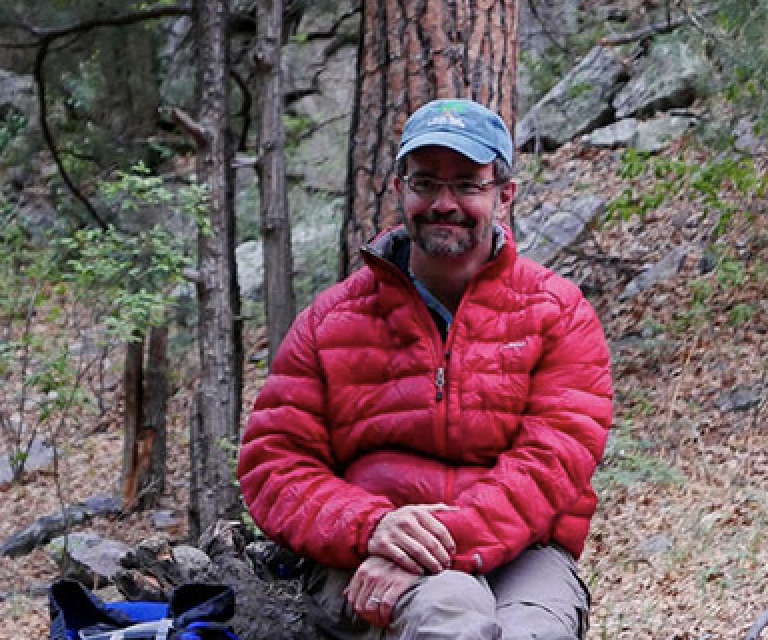 Stuart Harris, in a red puffy jacket and baseball cap sits by a tree in a rocky, mountainous spot.