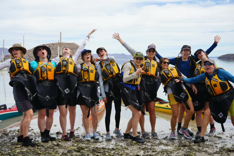 A group of women wearing sea kayak skirts and PDFs pose together in front of the ocean. They are standing on a beach and it's sunny. 