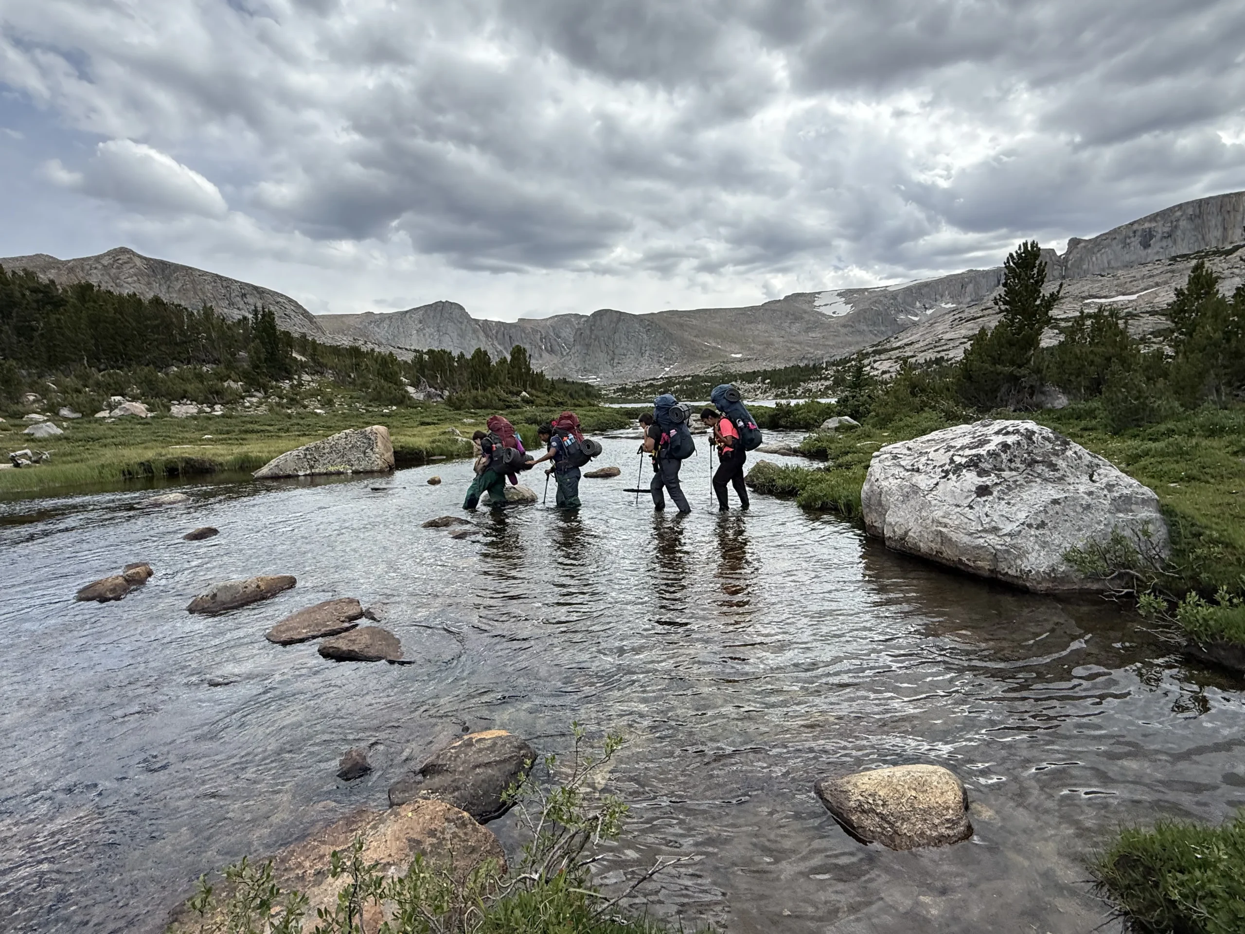 four people wearing large backpacks cross a small river. There are mountains in the distance. The sky is grey with clouds.