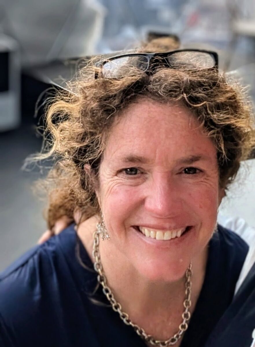 a woman with curly hairs in a ponytail and glasses on her head smiles at the camera.