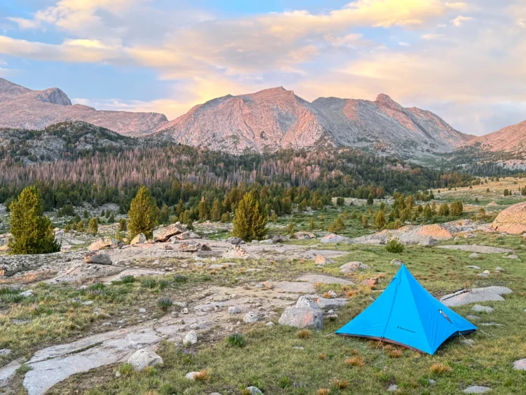 a blue tent sits in a rocky tundra area with mountains in the background