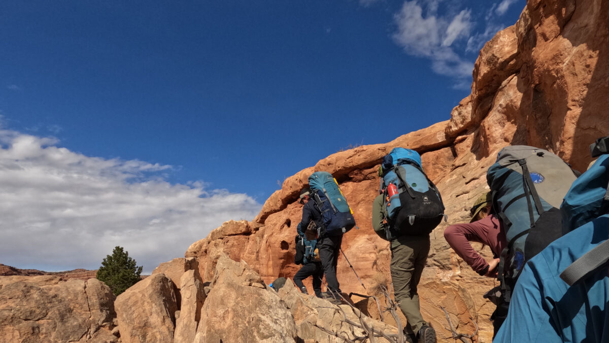 three people wearing large backpacking backpacks hike up a rocky trail of red rocks along a canyon wall