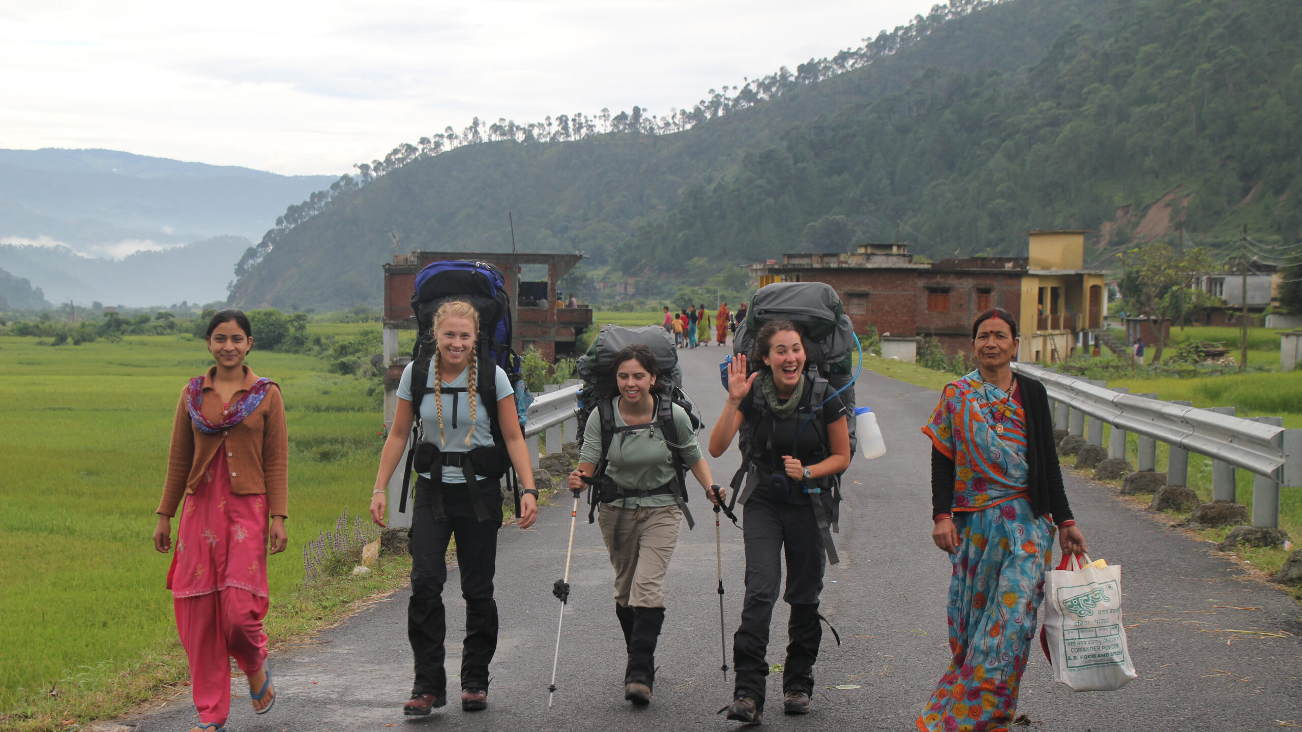 Three women with backpacks walk between two women wearing traditional Indian clothing.