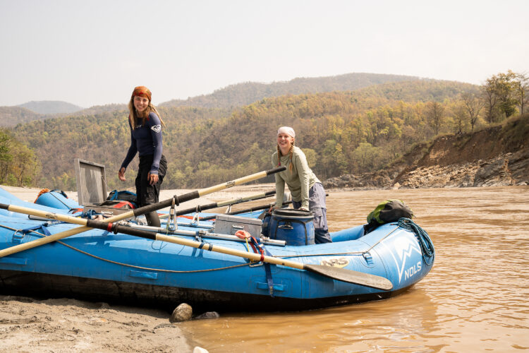 two women standing in a blue whitewater raft smile at the camera