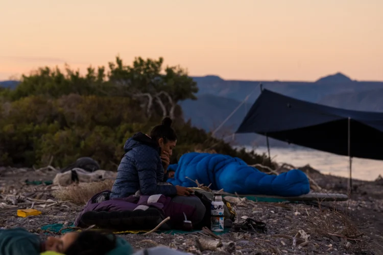 a person reads in a sleeping bag at dusk on a beach. there are people sleeping in sleeping bags on either side of them