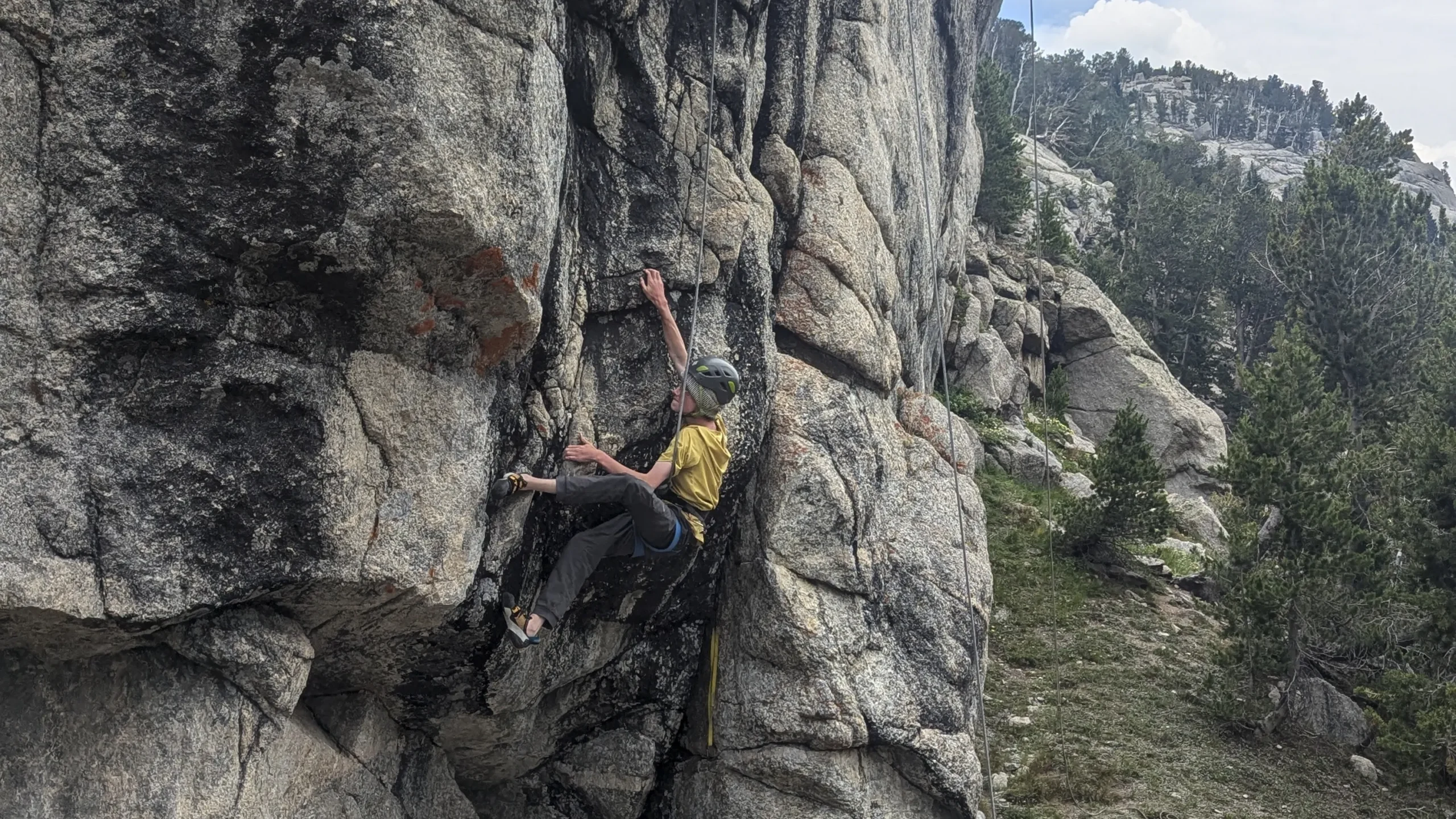 a person in a yellow shirt and a rock climbing helmet climbs a granite rock face. There are trees behind them.
