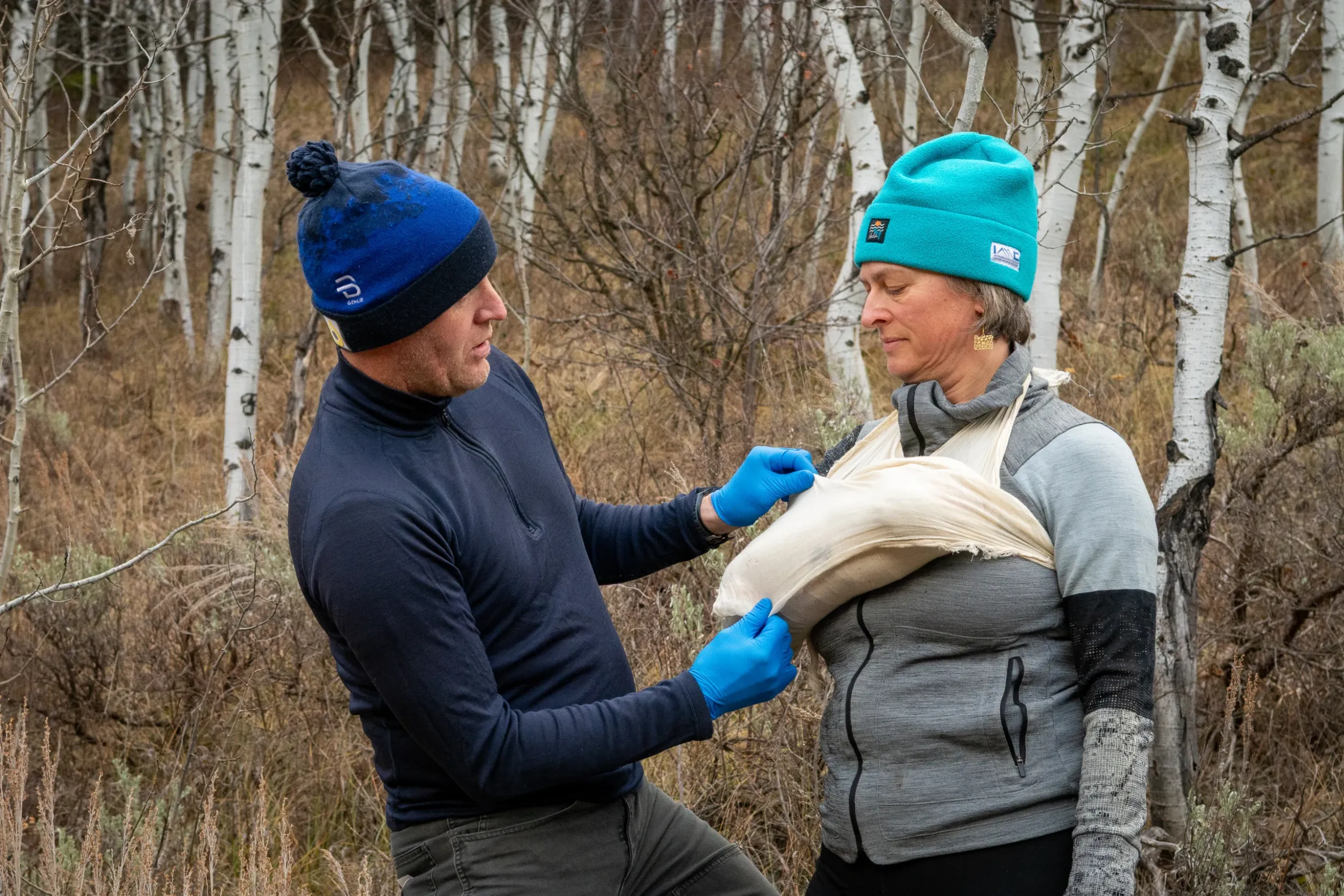 a woman in a blue beanie holds her arm in a sling while a man in a blue hat and blue latex gloves adjusts the sling