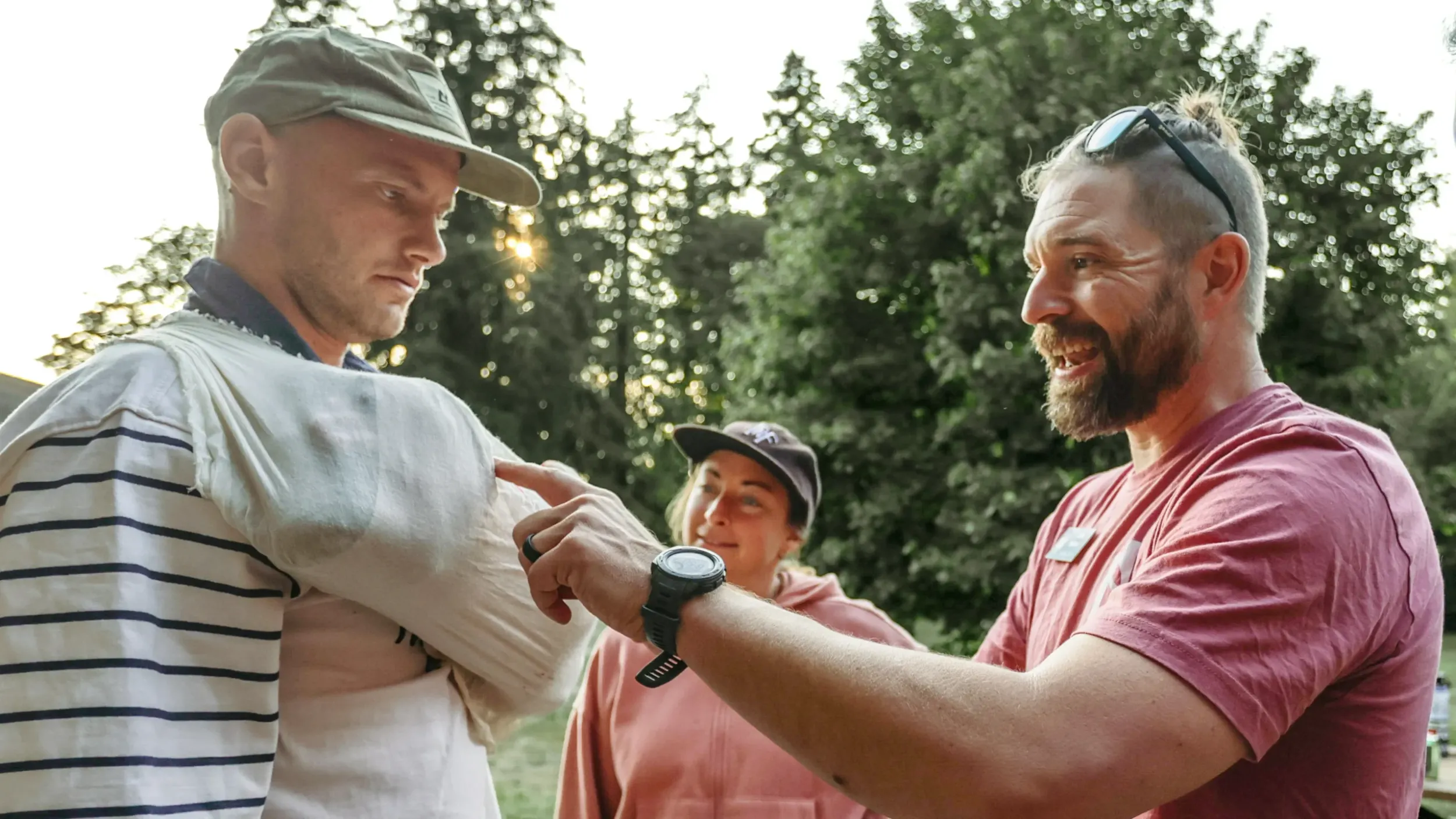 a man in a red shirt helps adjust a mock sling on the arm of another man.