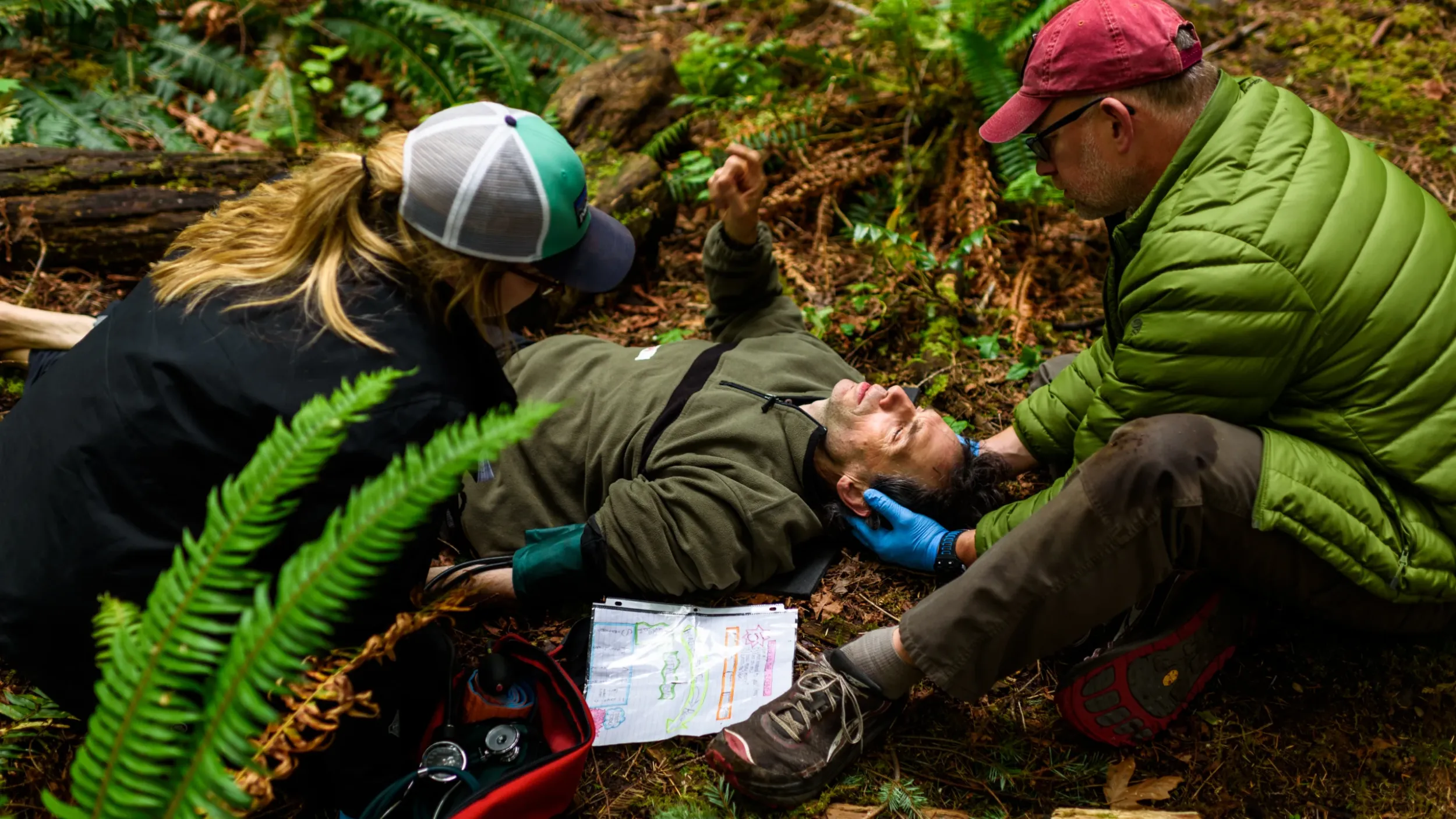a person wearing blue medical gloves holds a man's head. they are in the forest