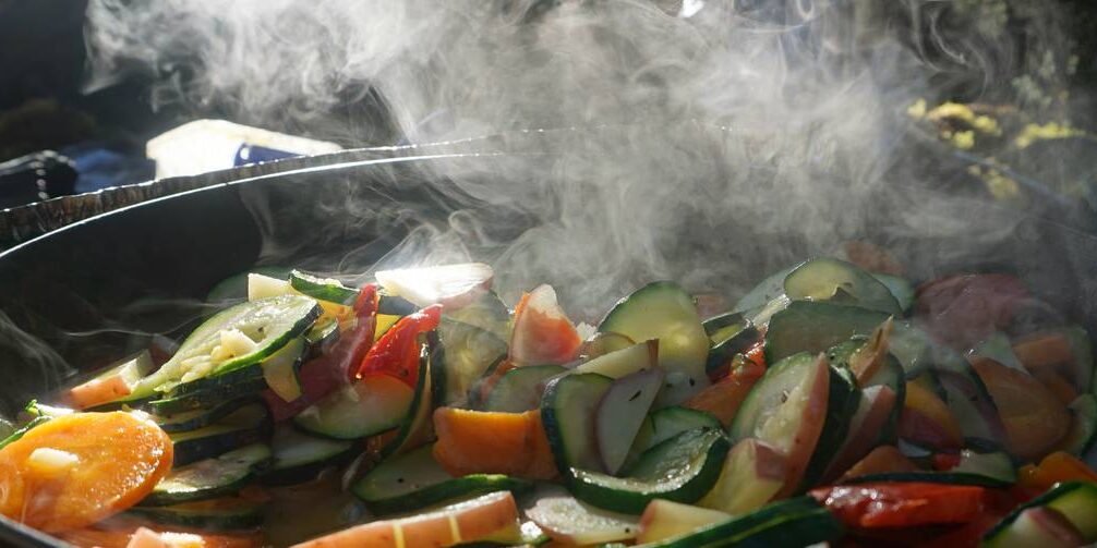 A pot of fresh cucumbers, carrots, peppers, and other vegetables steams over a camping stove.